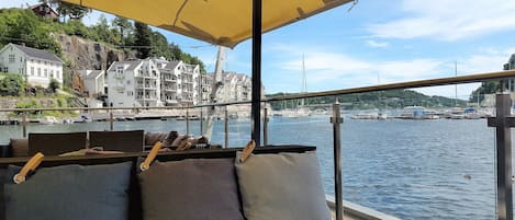 A boat deck with a yellow canopy, a seating area with cushions, and a view of a harbor with buildings and boats.