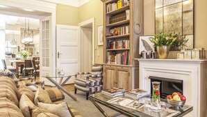 A living room with a sofa, armchair, fireplace, bookshelf, and dining area. - Hotel Elba am Kurfürstendamm - Design Chambers (Berlin)