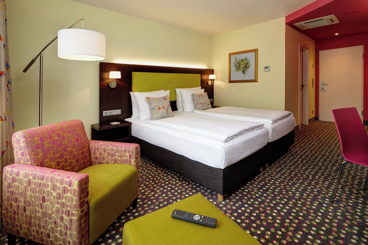 A hotel room with a bed, two nightstands, a lamp, a chair, and a remote control on a small table.
