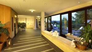 Un pasillo de hotel con pared de madera, plantas en macetas, un banco con toallas y vistas a la terraza exterior con sillas y una mesa.
