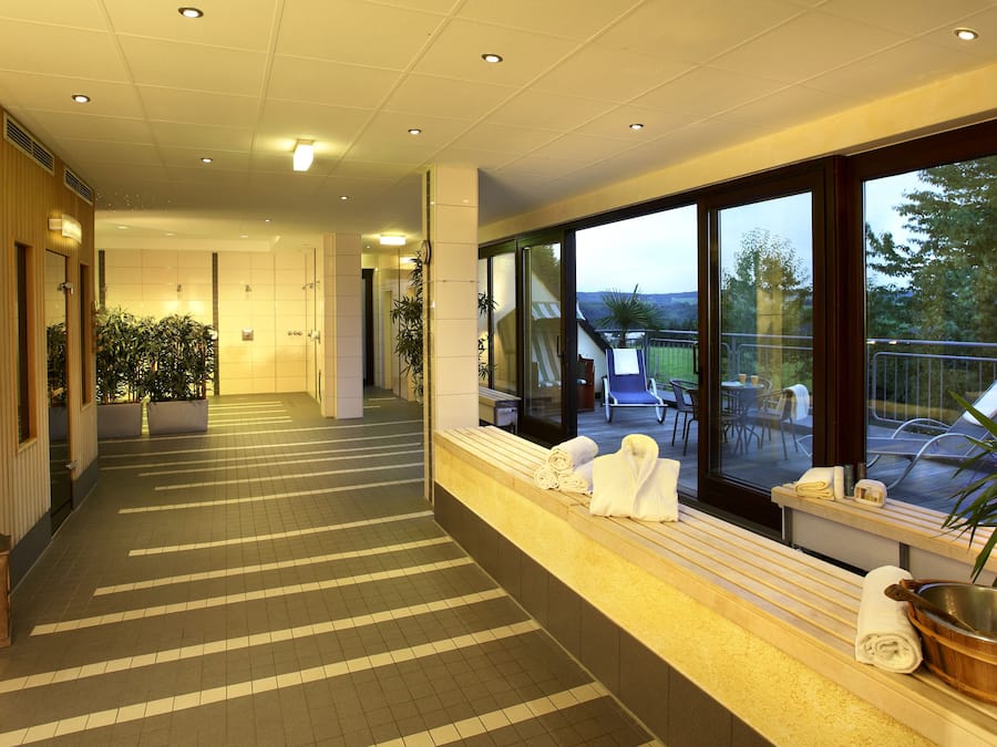 Un pasillo de hotel con pared de madera, plantas en macetas, un banco con toallas y vistas a la terraza exterior con sillas y una mesa.