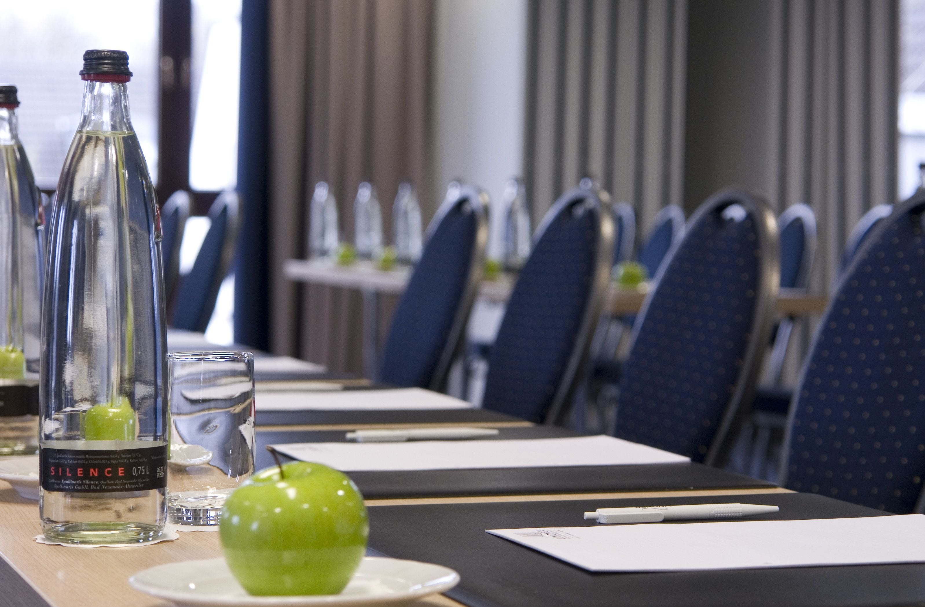 Et konferencelokale med et bord, stole, en flaske SILENCE 0.75L, et glas og et grønt æble.