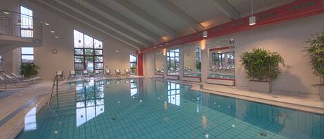 Una piscina interior con agua cristalina azul, ventanas iluminadas por el sol y sillones de descanso a los lados.