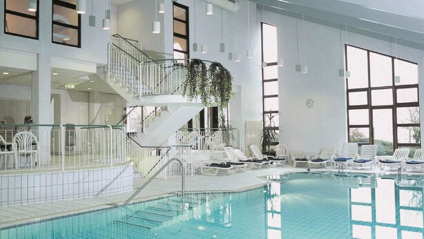 Una piscina interior con una escalera central, sillas de descanso y ventanas amplias.