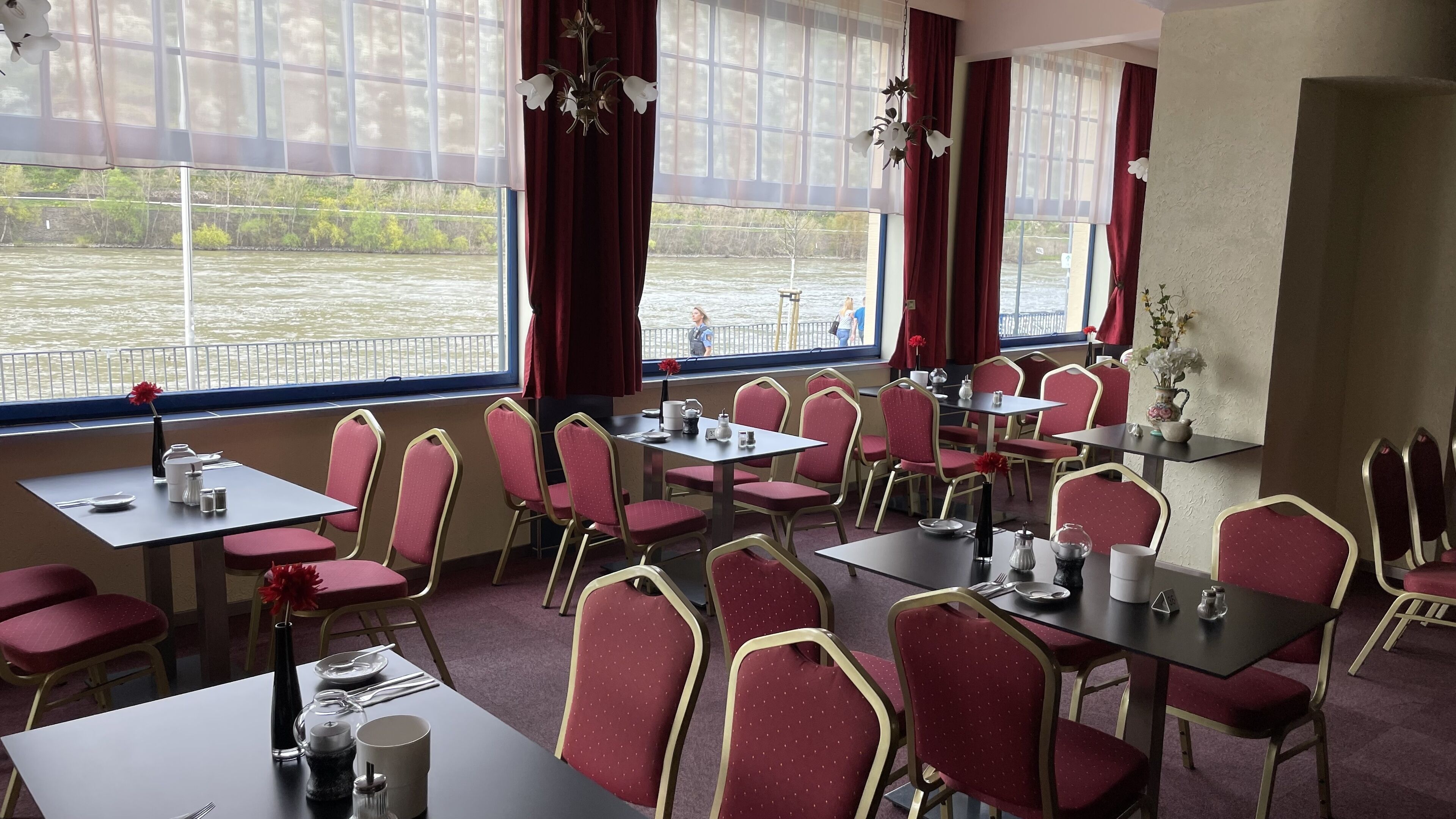 A dining area with tables and chairs, large windows with curtains, and a view of a body of water.