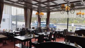 A restaurant with tables and chairs, large windows with curtains, and a view of boats on a river. - Hotel Rheinlust (Boppard)