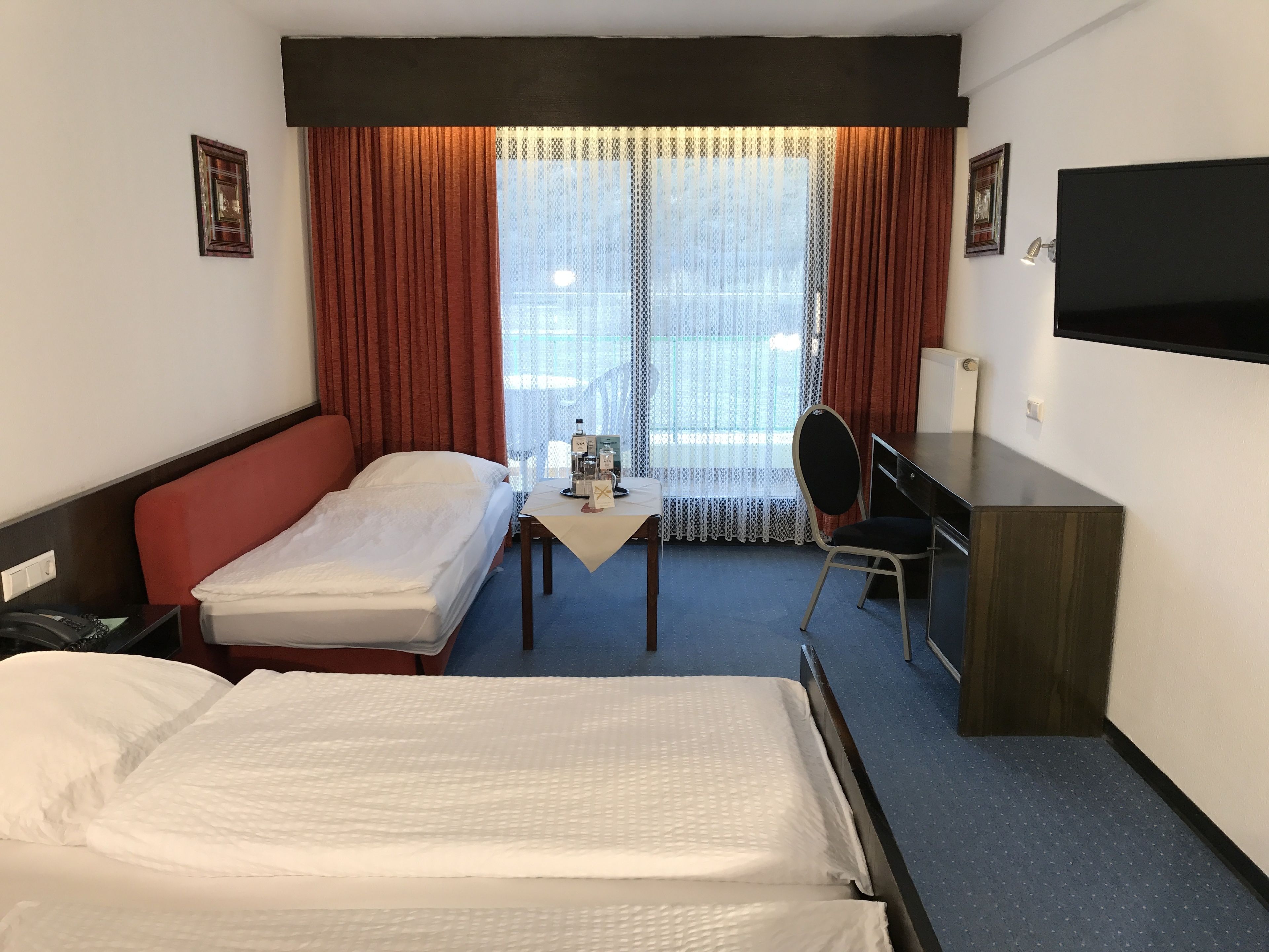 Habitación de hotel con una cama grande, una cama más pequeña, un escritorio, una silla, un televisor y una ventana con cortinas.