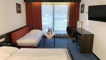 Habitación de hotel con una cama grande, una cama más pequeña, un escritorio, una silla, un televisor y una ventana con cortinas.