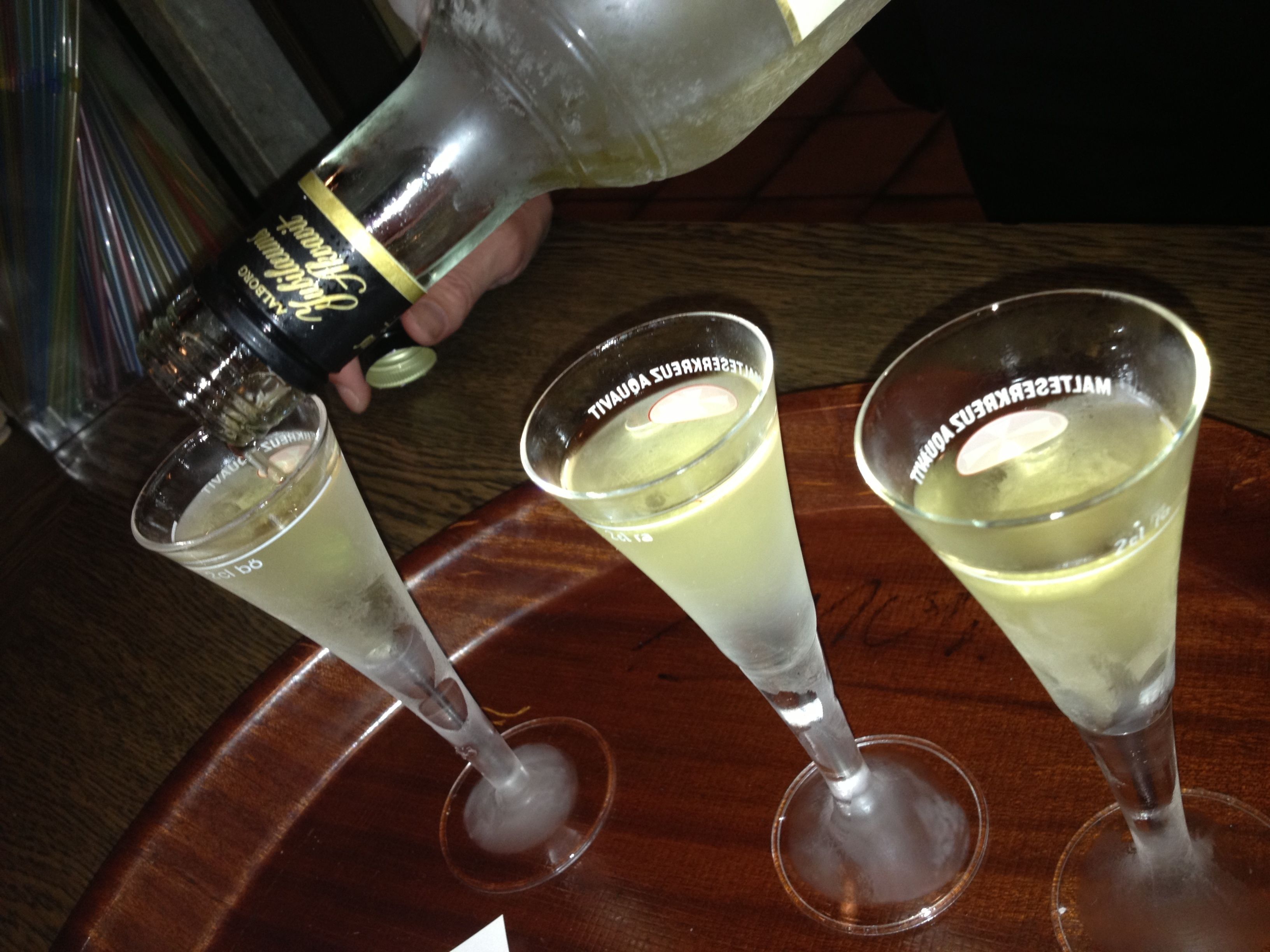 Three glasses on a tray, each with a label reading "TIVAUX". A hand is pouring a clear liquid into one of the glasses.