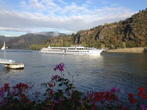 Un gran barco de crucero fluvial llamado 'Staten Island' navegando sobre un río tranquilo, con un muelle en primer plano y una ladera boscosa al fondo.