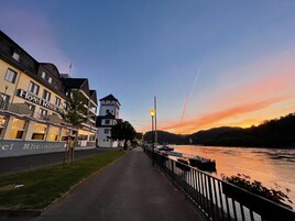 Un paseo fluvial con un hotel llamado 'Hotel Rheinlust' a la izquierda, con balcones y una torre.