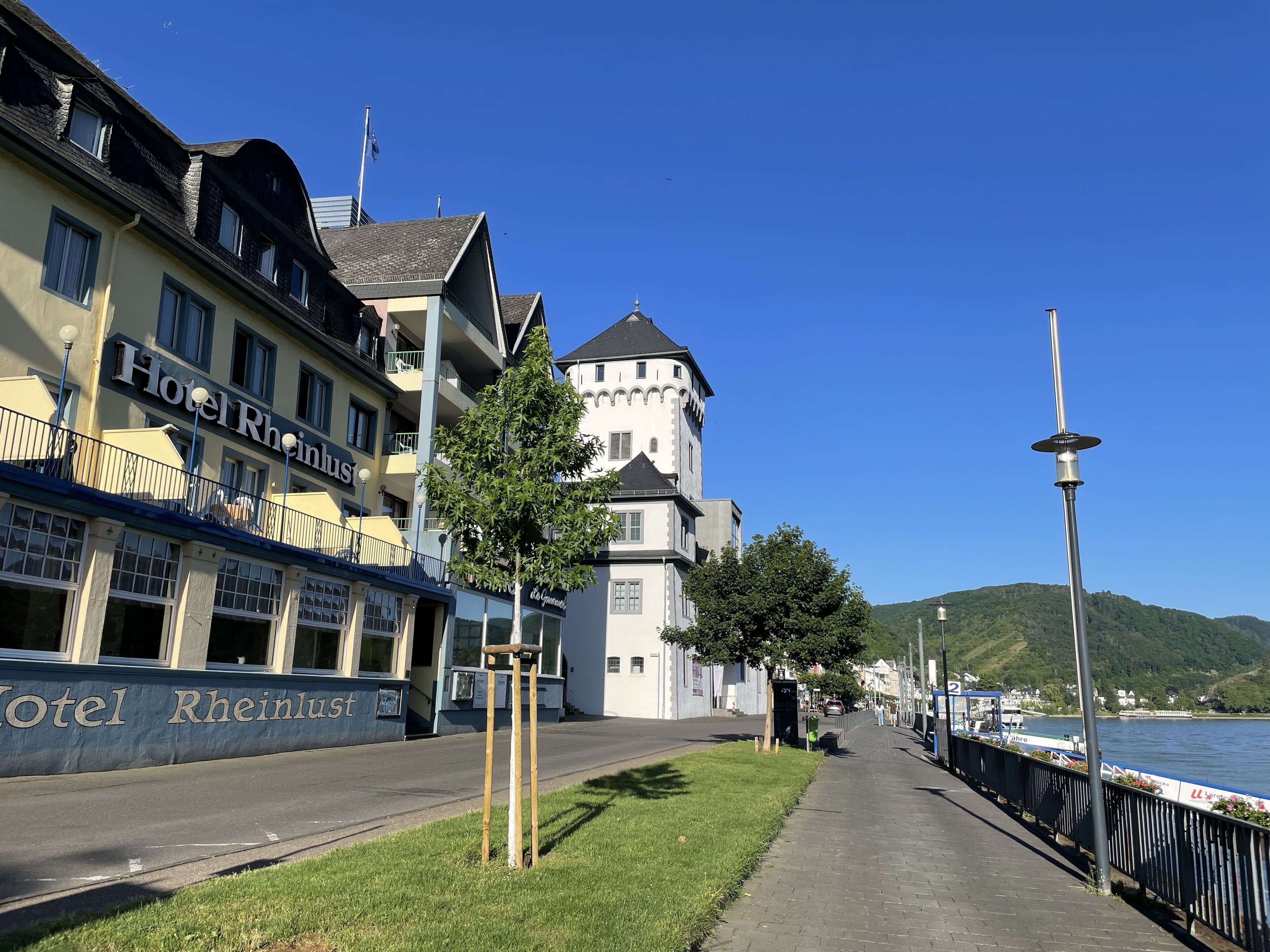 A hotel named 'Hotel Rheinlust' with a traditional building and a tower, located beside a river with a walkway and a fence.