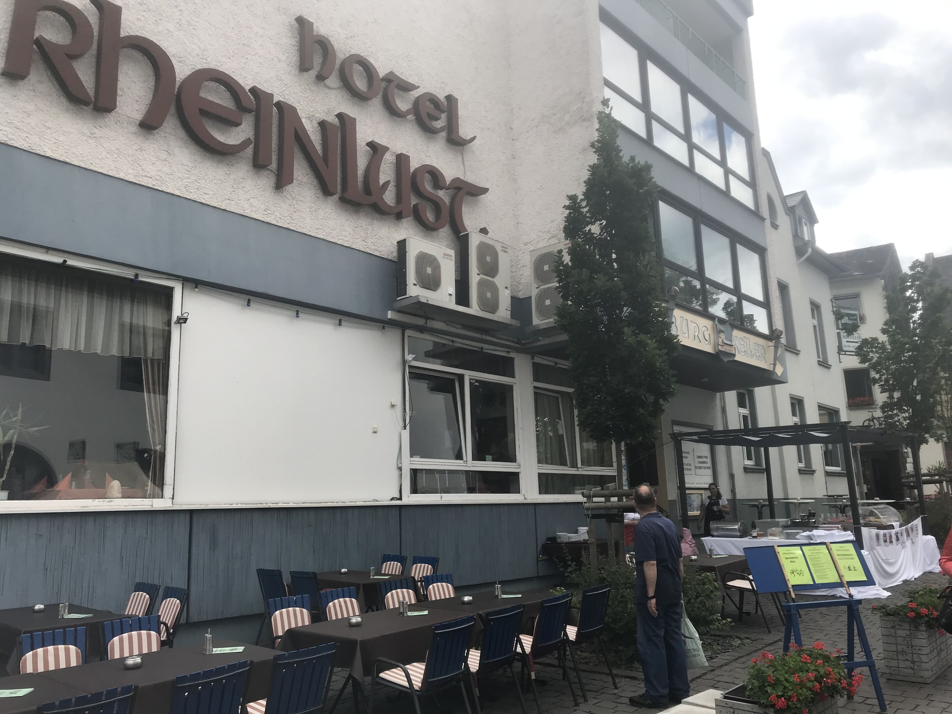 A hotel exterior with outdoor seating, a sign reading "hotel Rheinlust," and a person standing near the entrance.