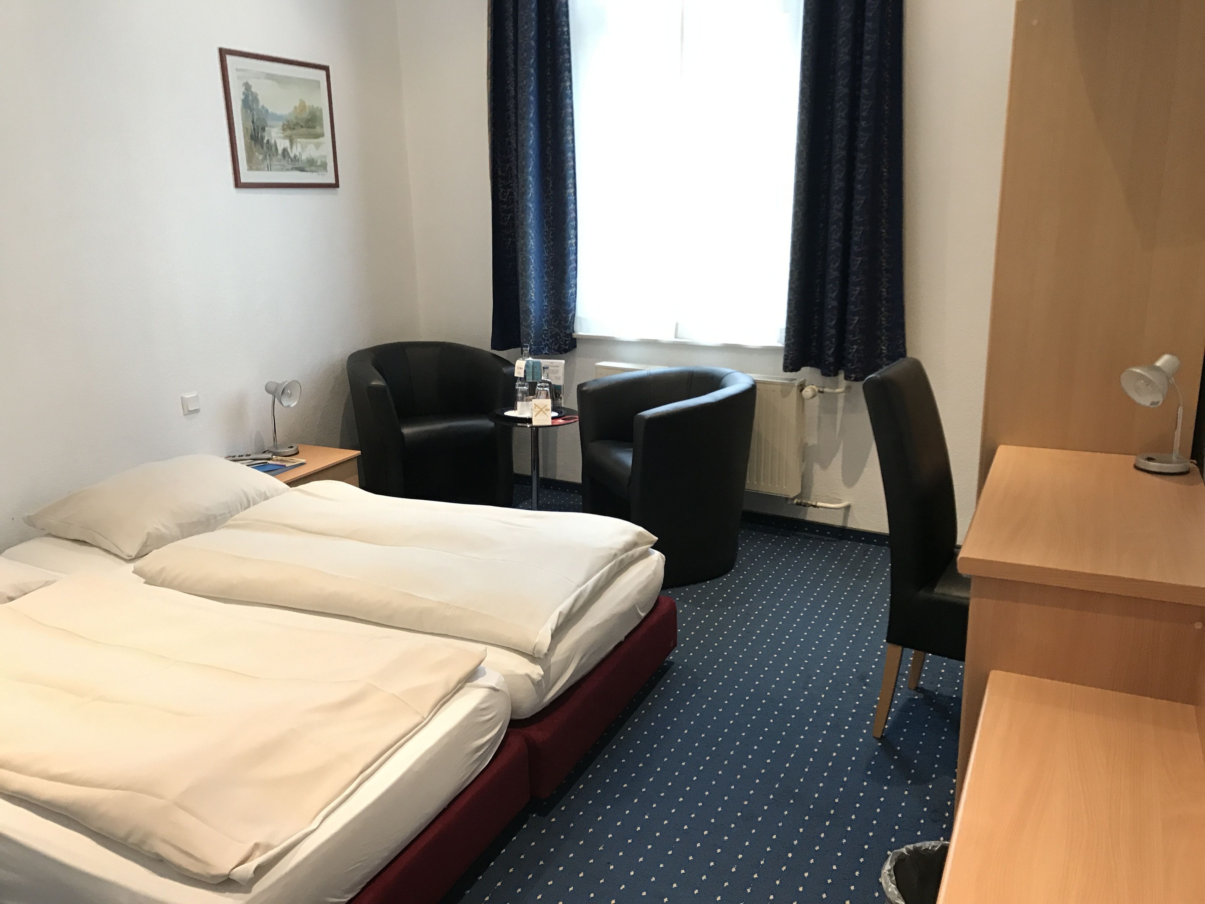 Ein Hotelzimmer mit Bett, zwei Stühlen, Schreibtisch, Lampe und einem Fenster mit Vorhängen.
