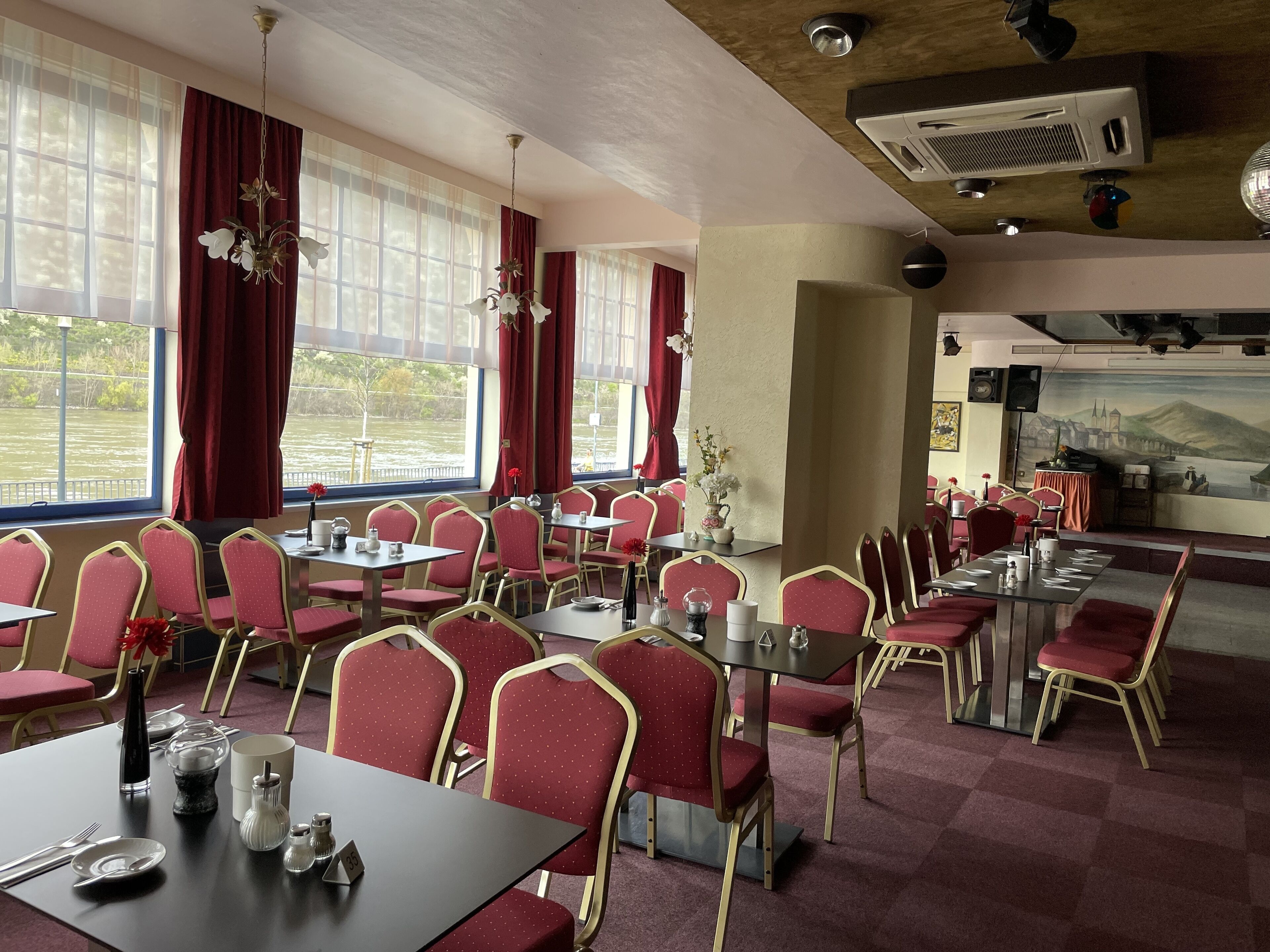 Un restaurant avec des chaises et des tables rouges, de grandes fenêtres avec des rideaux et une fresque murale.