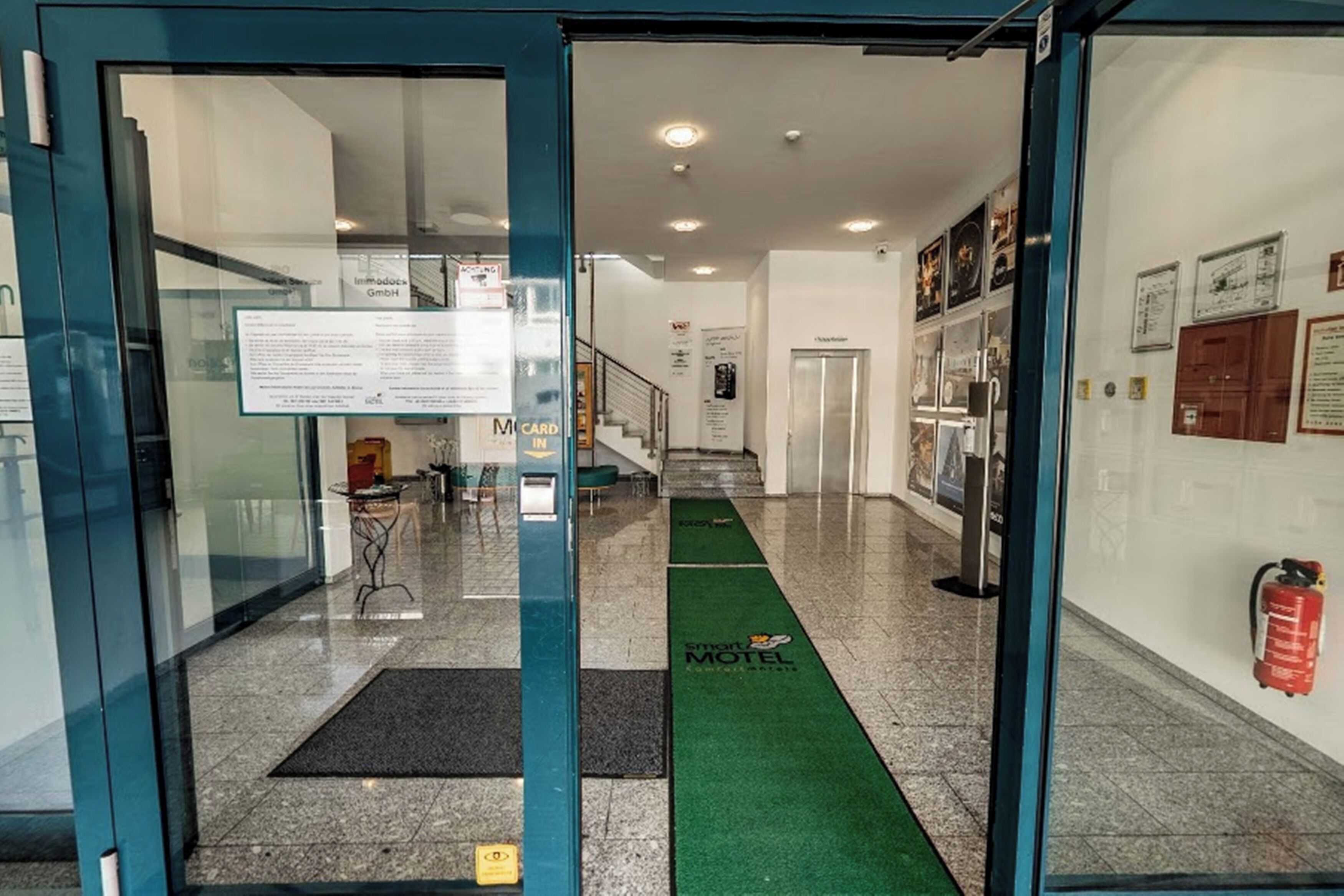 A hotel entrance with a green carpet, a reception desk, and a staircase leading to the upper floor.