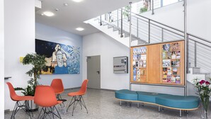 Un vestíbulo con mostrador de recepción, sillas naranjas, un banco azul, una pared con un cuadro, una escalera y un tablón de anuncios.