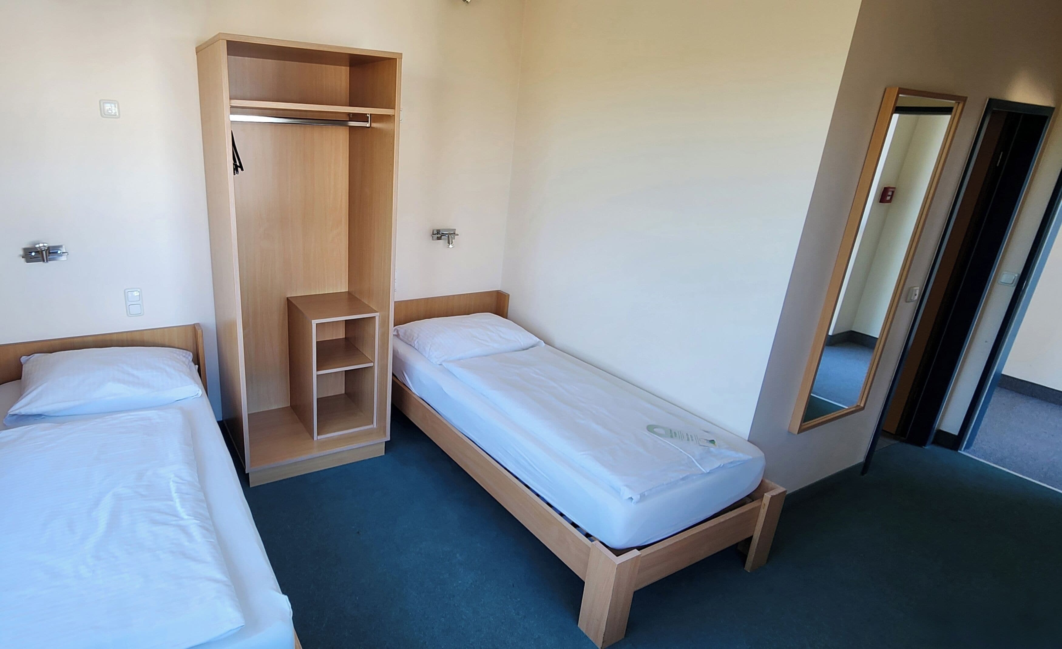 A small, single-room accommodation with two single beds, a wooden wardrobe, and a mirror.