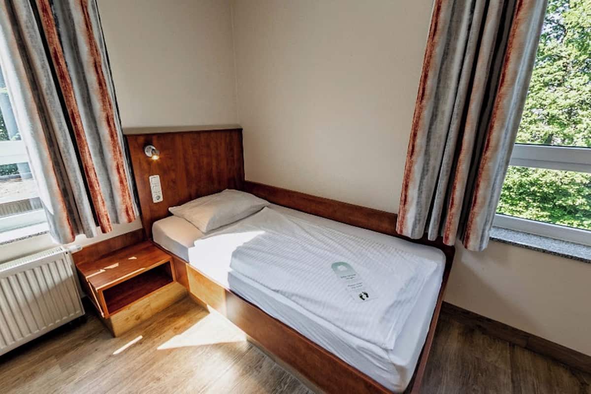 A small, single-bed room with a wooden headboard, a bedside table, and a window with curtains.