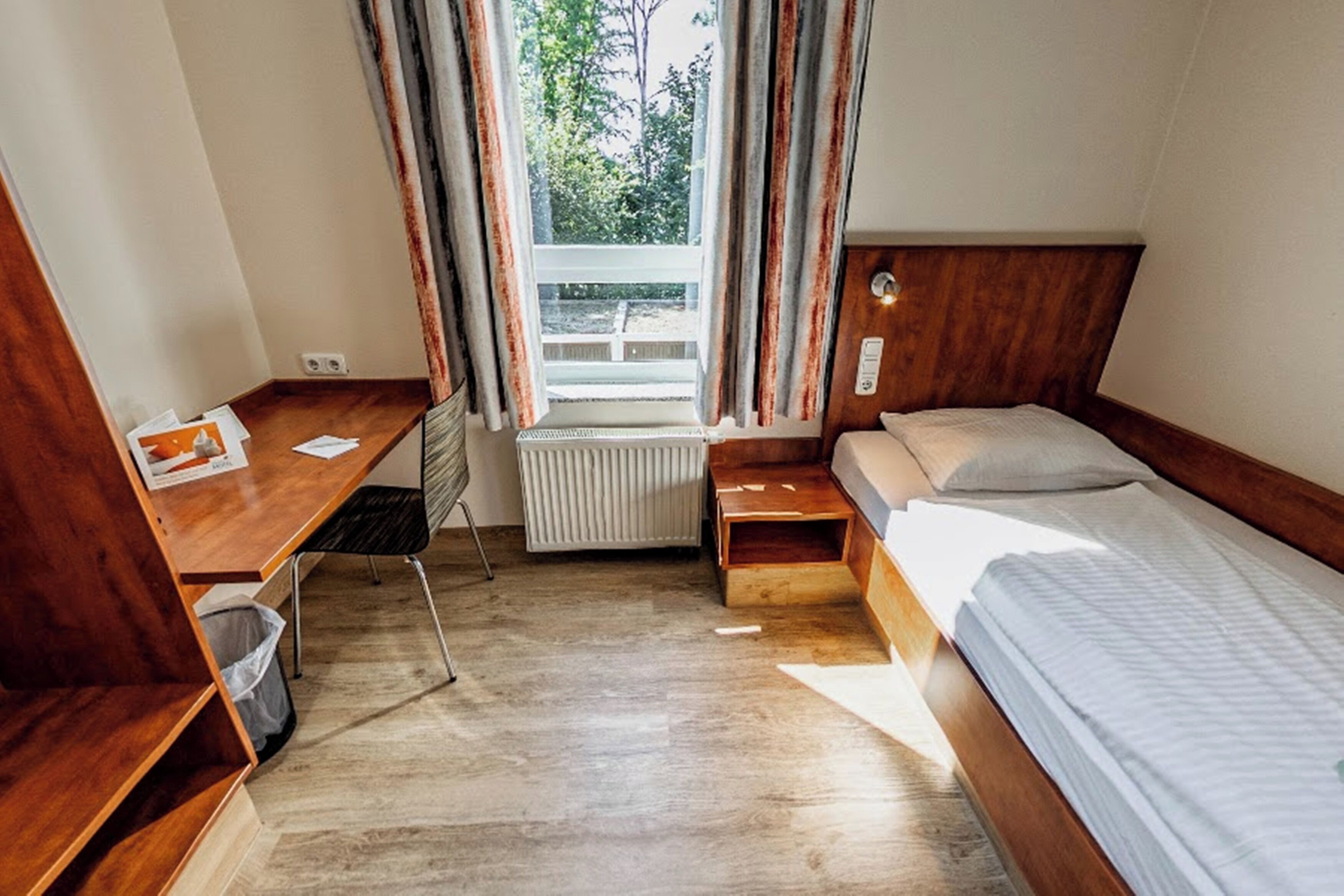 A compact room with a wooden bed, a small desk, a radiator, and a window with curtains.