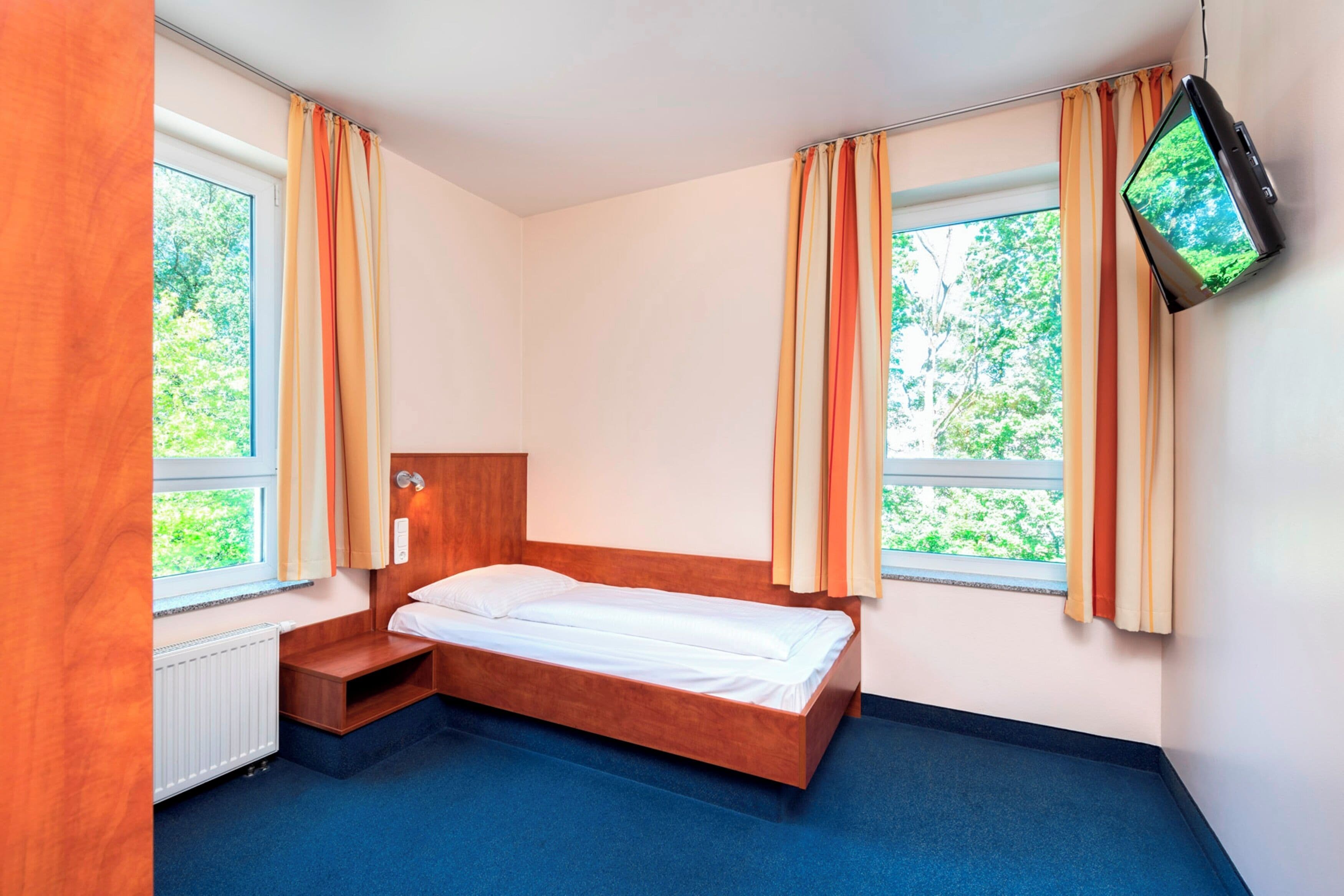 A small, single-bed room with a wooden headboard, a TV mounted on the wall, and large windows with curtains.