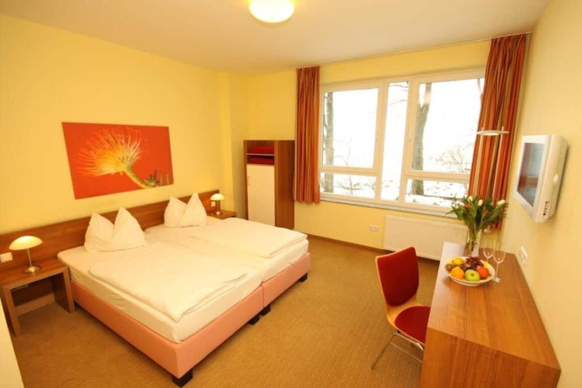 A hotel room with a double bed, a desk with a chair, a TV, a window with curtains, and a fruit bowl on the desk.