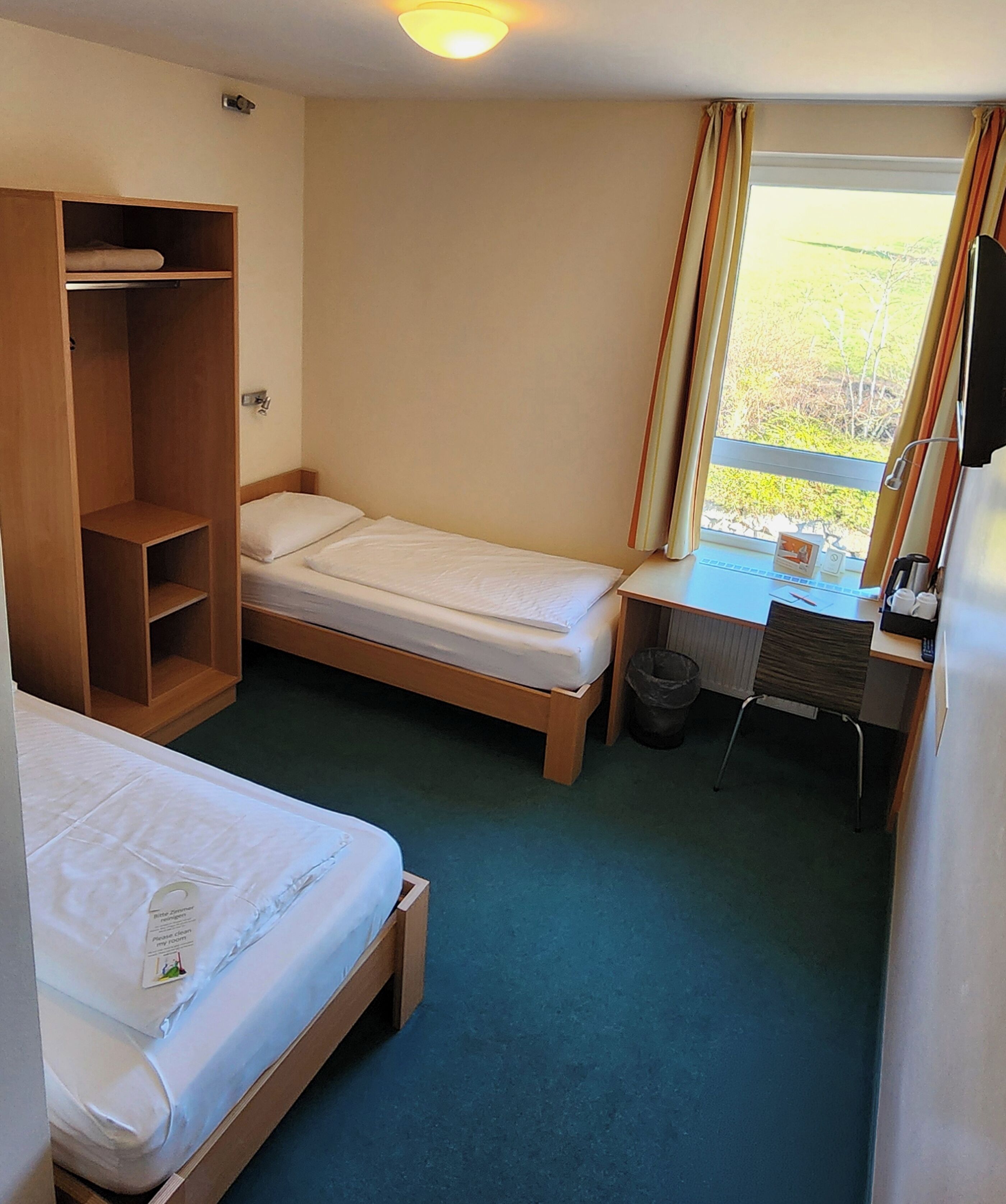 Quarto de hotel pequeno com duas camas de solteiro, um guarda-roupa de madeira, uma escrivaninha com cadeira e uma janela com cortinas.