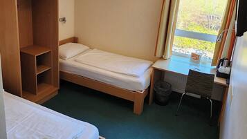 Quarto de hotel pequeno com duas camas de solteiro, um guarda-roupa de madeira, uma escrivaninha com cadeira e uma janela com cortinas.