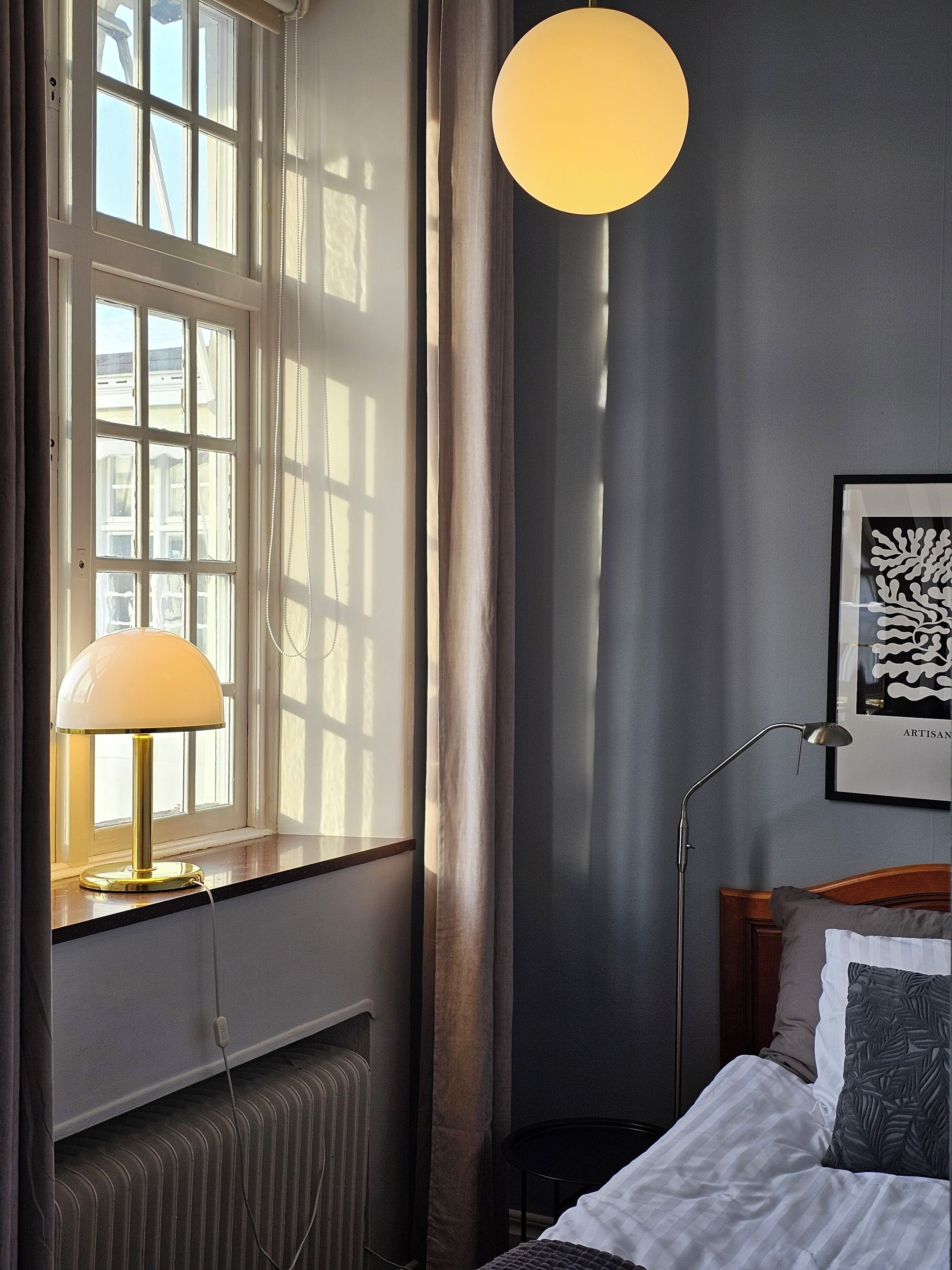 A bedroom with a bed, a bedside lamp, a radiator, a window with curtains, and a wall-mounted light fixture.