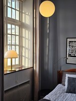 Un dormitorio con cama, lámpara de noche, radiador, ventana con cortinas y aplique de pared.