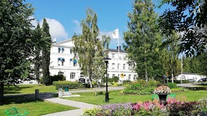 A park with a green bench, a pathway, and a white building with a flag. - Furunäset Hotell och Konferens (Pitea)