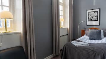 Quarto de hotel com cama, duas luminárias de cabeceira, radiador, uma obra de arte emoldurada na parede e janela com cortinas.