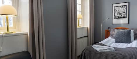Una habitación de hotel con cama, dos lámparas de noche, un radiador, una obra de arte enmarcada en la pared y una ventana con cortinas.