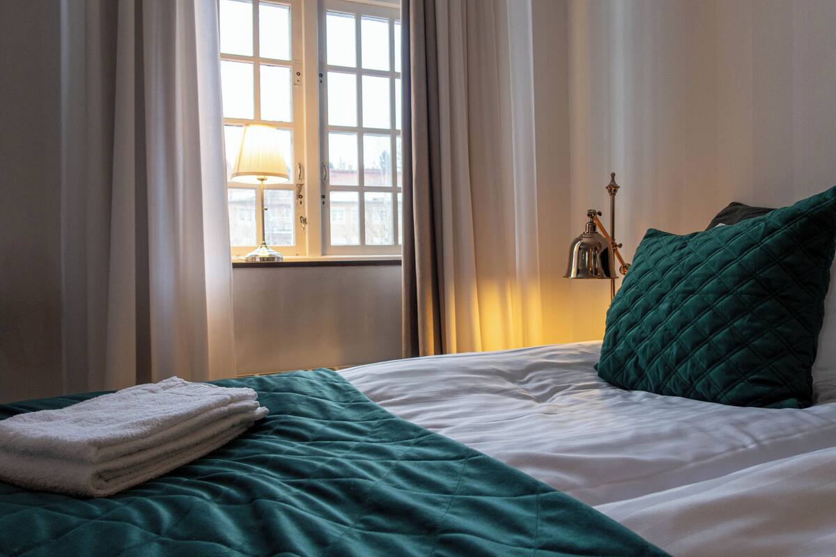 A neatly made bed with a teal bedspread and a white blanket, a window with curtains, and a wall-mounted lamp.
