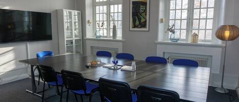 A conference room with a long table, blue chairs, a white cabinet, a painting, and a window with a view.