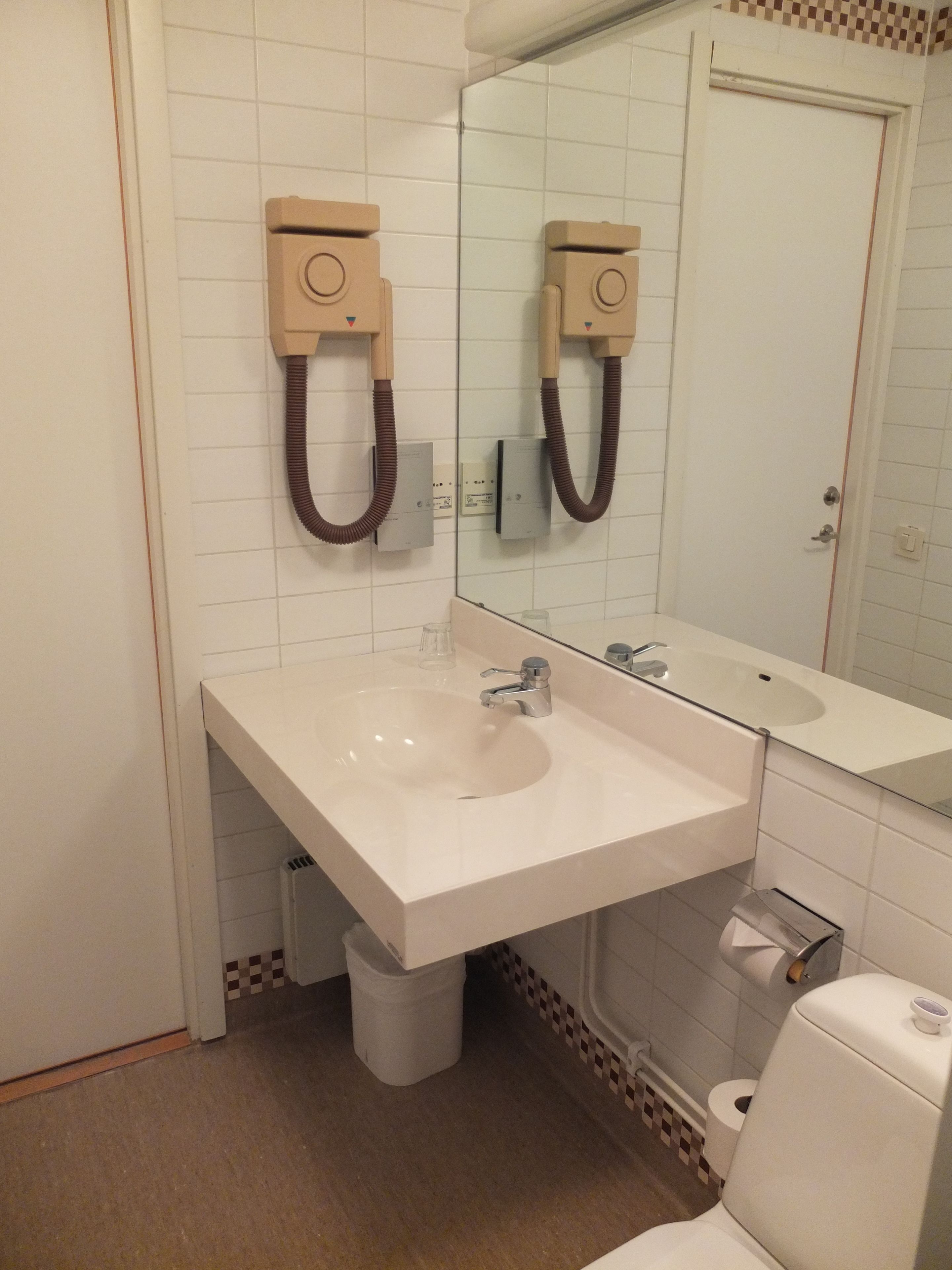 A bathroom with a white sink, a mirror, and a toilet. There is a trash can and a paper towel dispenser.
