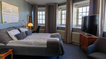 Ett hotellrum med en stor säng, ett skrivbord med en lampa, en TV och fönster med gardiner.