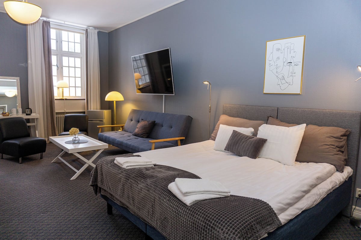 A hotel room with a large bed, a sofa, a desk, a TV, and a painting on the wall.