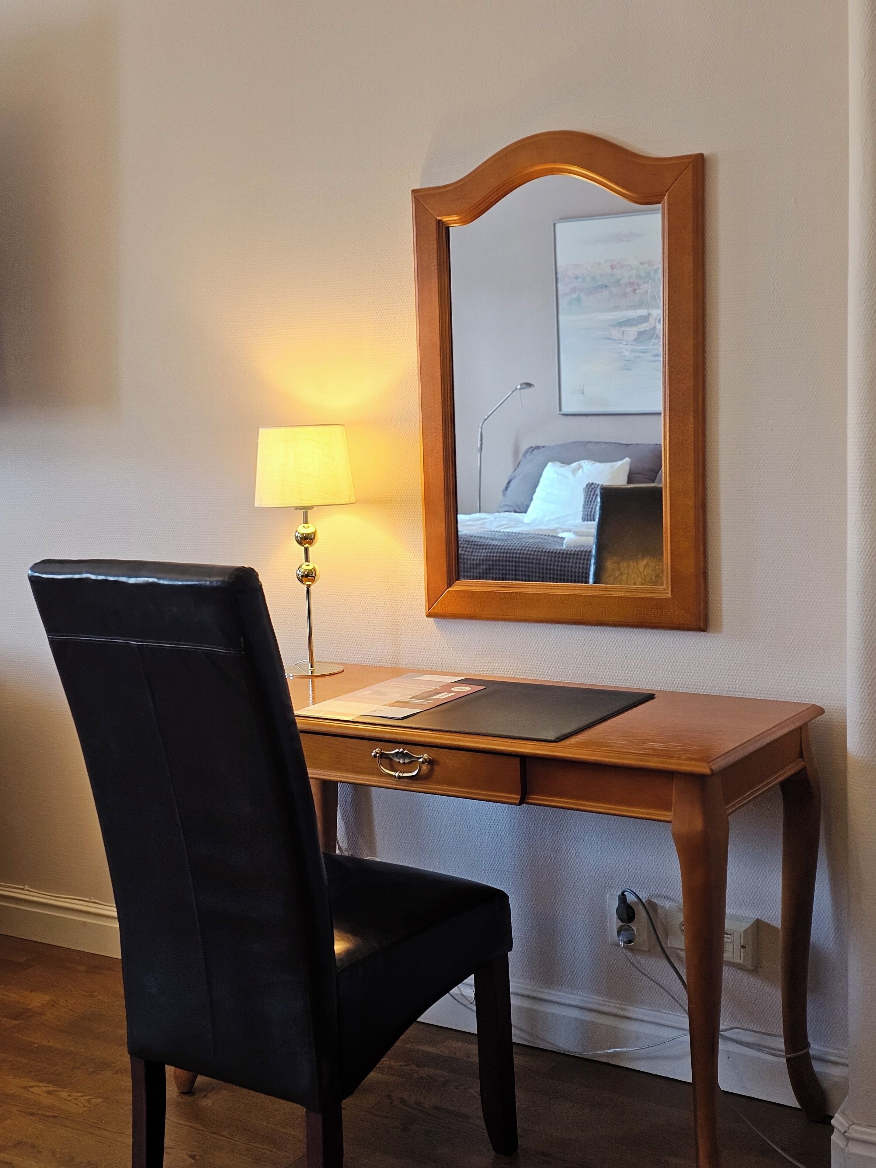 A wooden desk with a black chair, a lamp, and a mirror reflecting a bed and a painting on the wall.