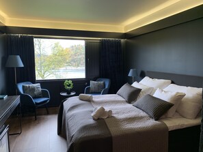 Quarto de hotel com uma cama grande, duas cadeiras, uma escrivaninha e uma janela com cortinas.
