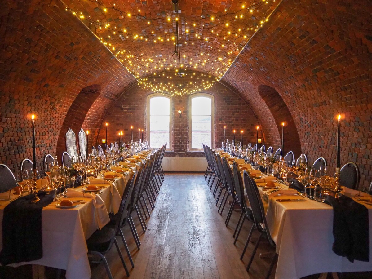 Curved, red brick room strung with lights across the ceiling. 2 long tables are set in the room for a fancy dinner
