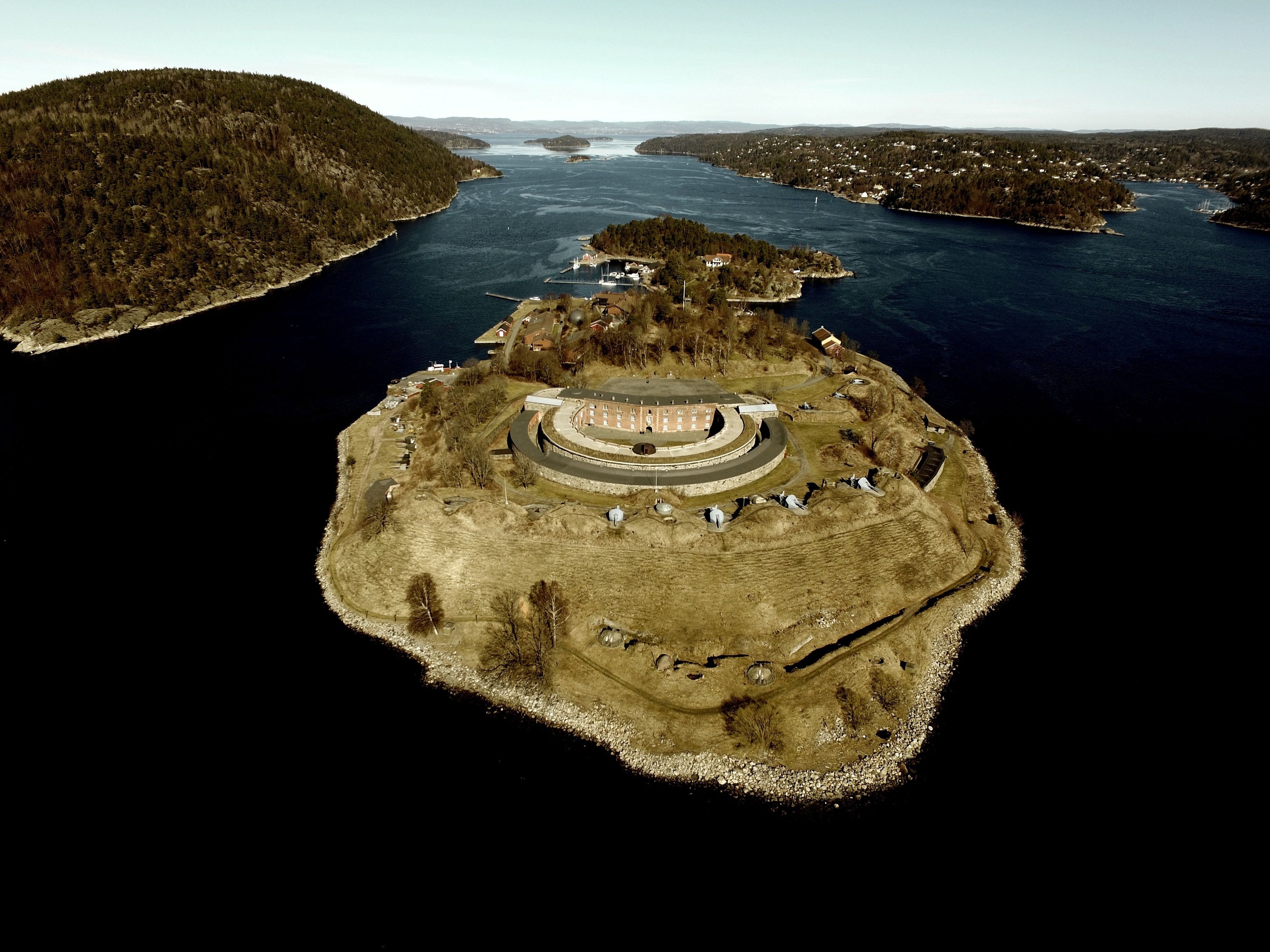 Ett flygbildsbild av en ö vid kusten med ett cirkelformat byggnadskomplex, omgivet av vatten och skogsområden.