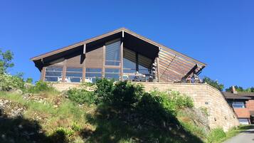 Un bâtiment moderne doté de grandes fenêtres et d’un toit en pente, situé sur une colline et entouré de verdure.