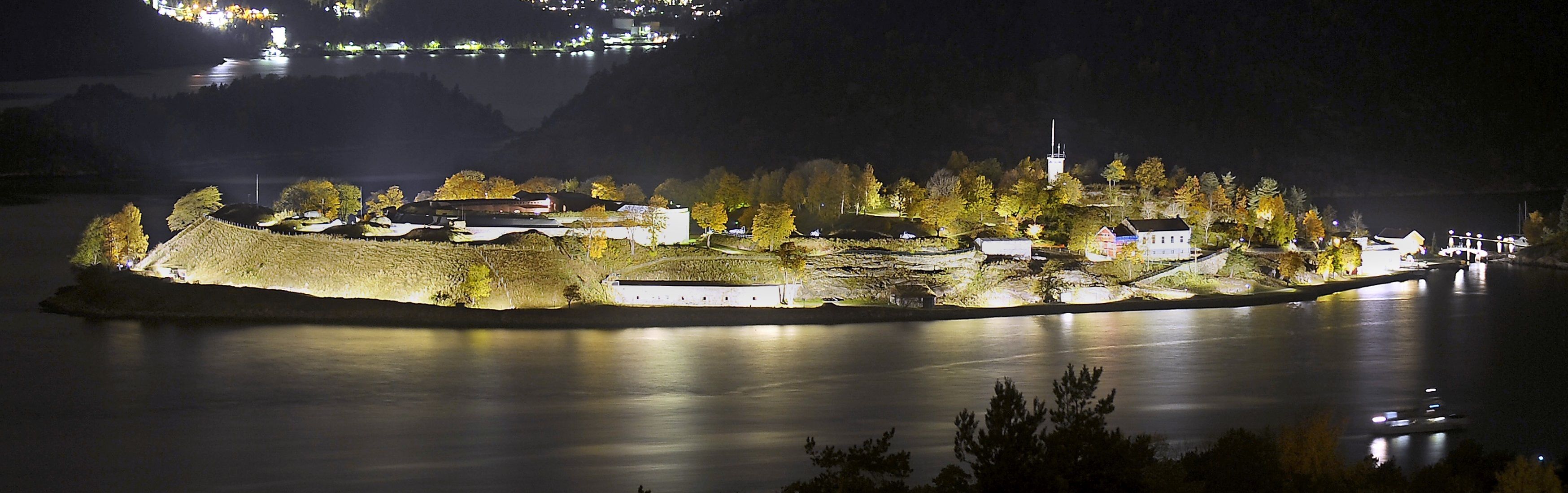A well-lit island with buildings and a dock, surrounded by calm water and trees.