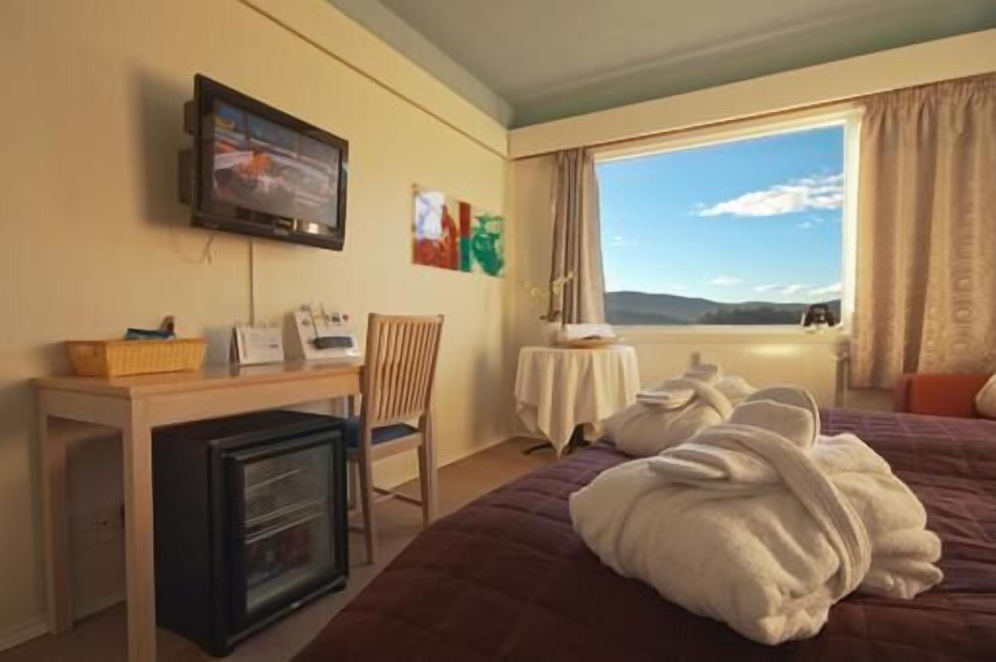 Quarto de hotel com cama, escrivaninha com televisão, mesa pequena e janela com vista.