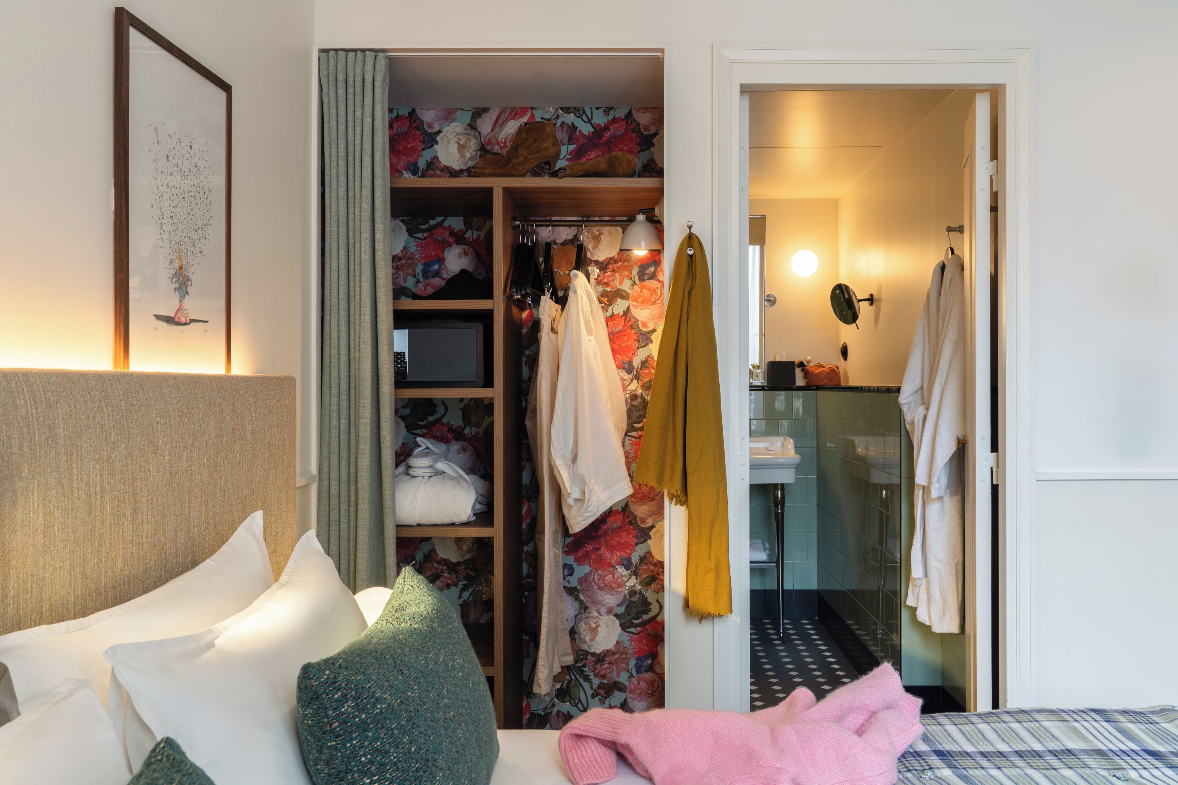 A hotel room with a bed, a closet with hanging clothes, a bathroom with a mirror, and a shower.