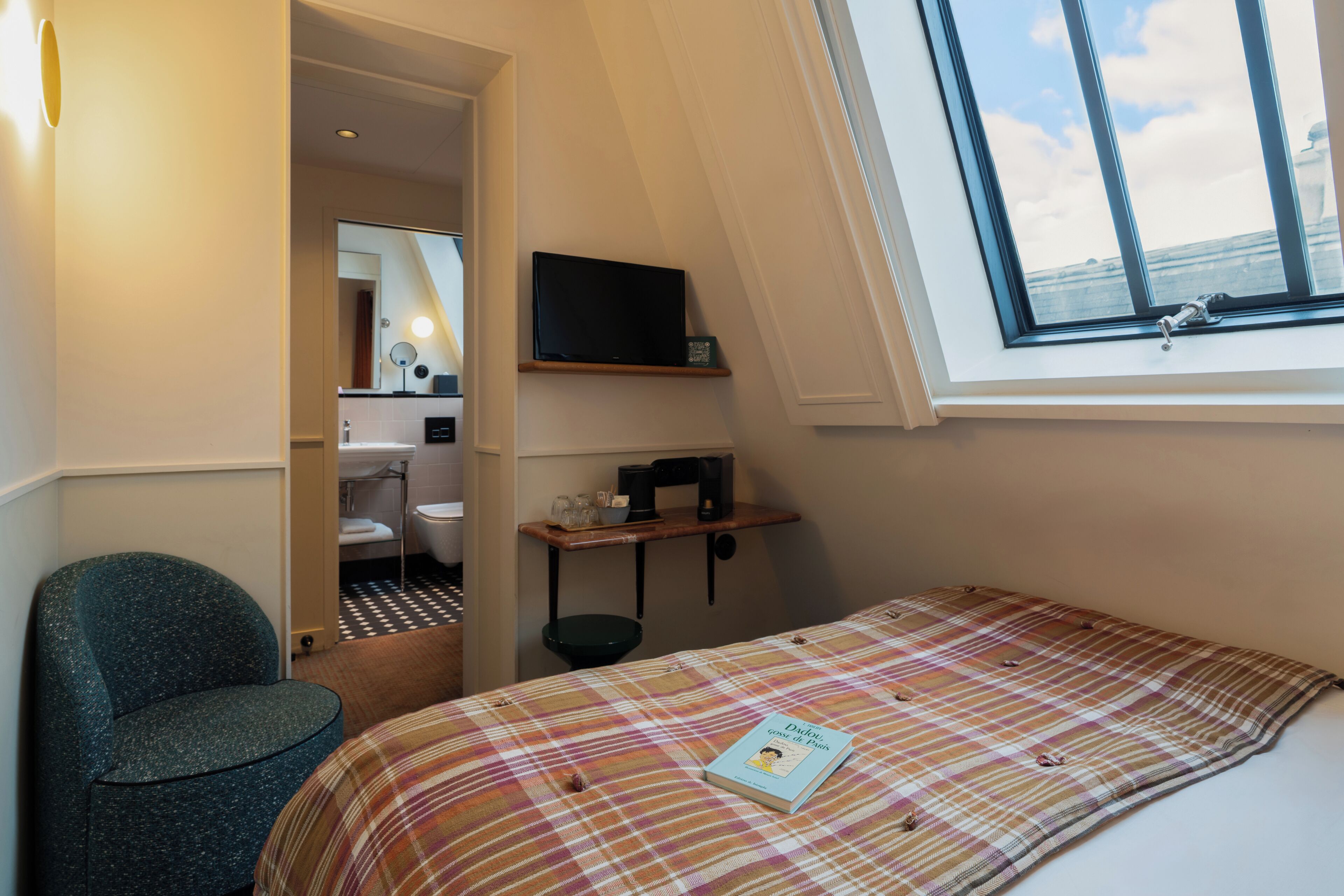 Una camera da letto con un letto, una sedia, una finestra, una TV, uno scaffale e un bagno visibile attraverso una porta aperta.