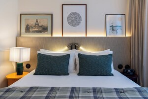 Habitación de hotel con cama, dos almohadas, mesita de noche con lámpara y cuadros en la pared.
