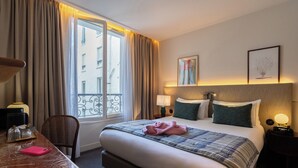 A hotel room with a bed, bedside tables, a desk, a chair, a window with curtains, and a view of buildings outside. - Dadou Paris (Paris)