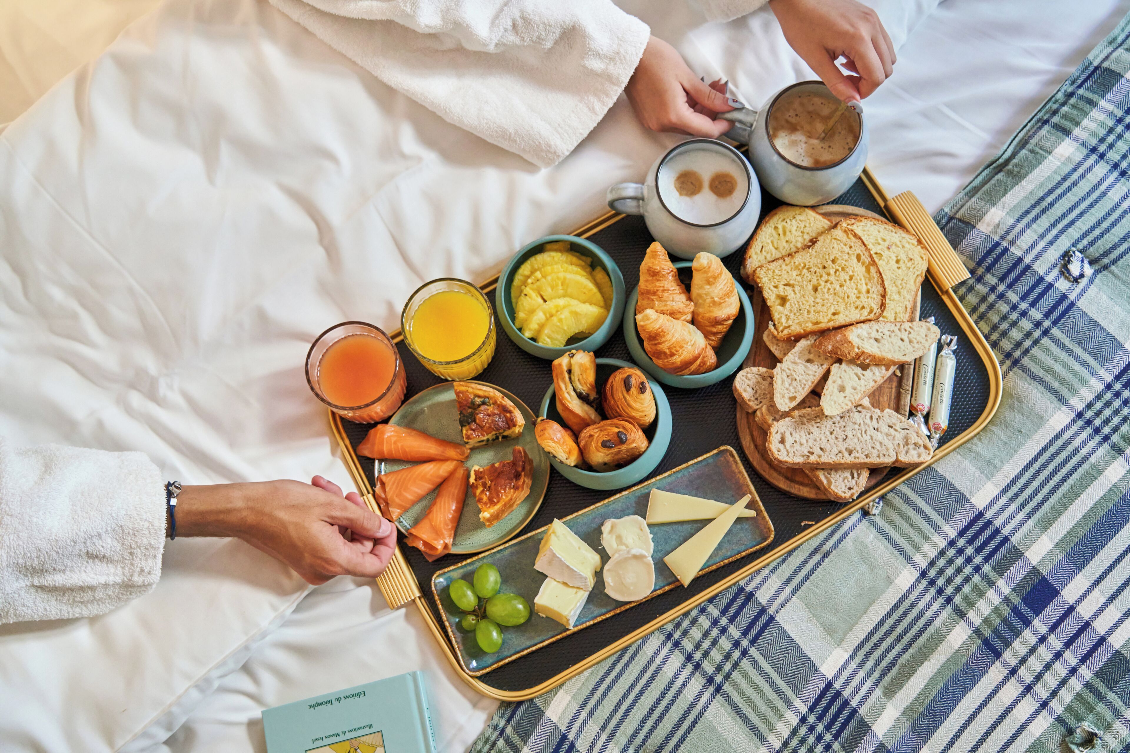 A breakfast tray with croissants, cheese, grapes, and a coffee mug on a bed with a checkered blanket.
